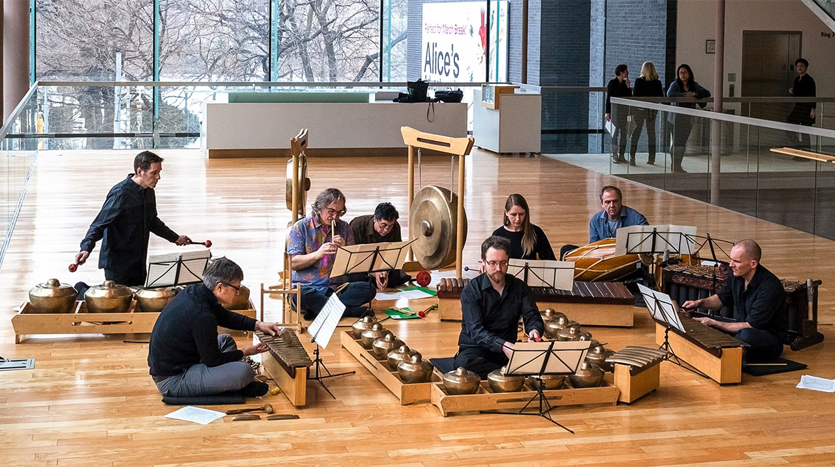 Sitting in a bright atrium, a group of musicians plays bronze instruments including flutes, gongs, and drums